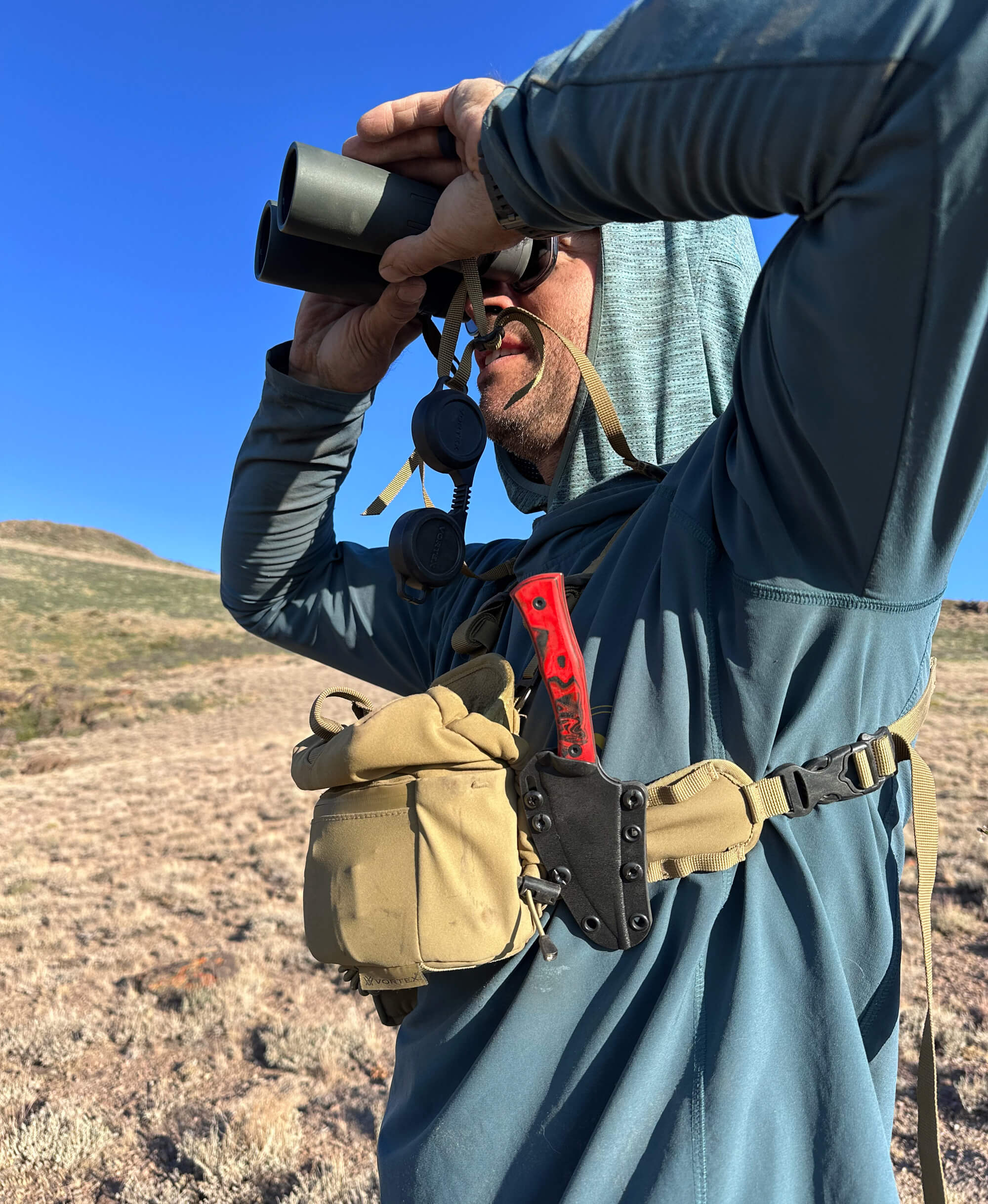 Person in outdoor setting using binoculars with a blue sky and landscape in the background. The person is shown with a red and black hunting knife in a black sheath.