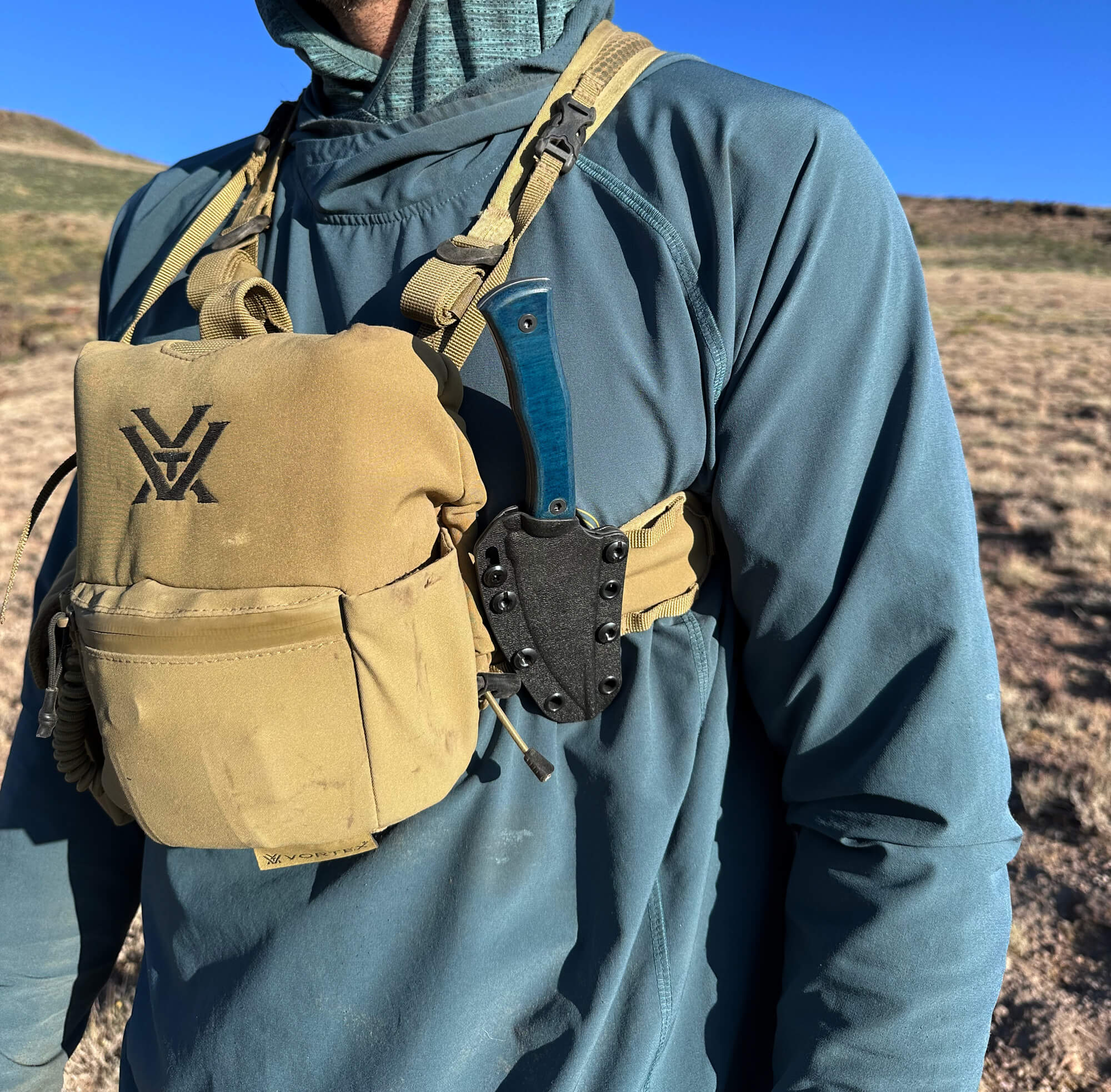 Person in outdoor setting with a blue sky and landscape in the background. The person is shown with a blue hunting knife in a black sheath.